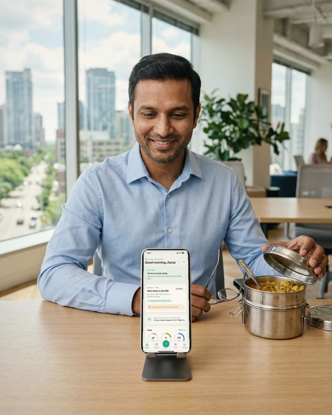 A man at an office desk abroad opens a steel tiffin, his phone on a stand showing his day in Sthira.